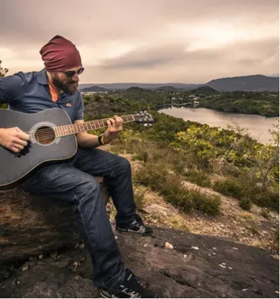 A sitting down and playing guitar with a mountainous background.
